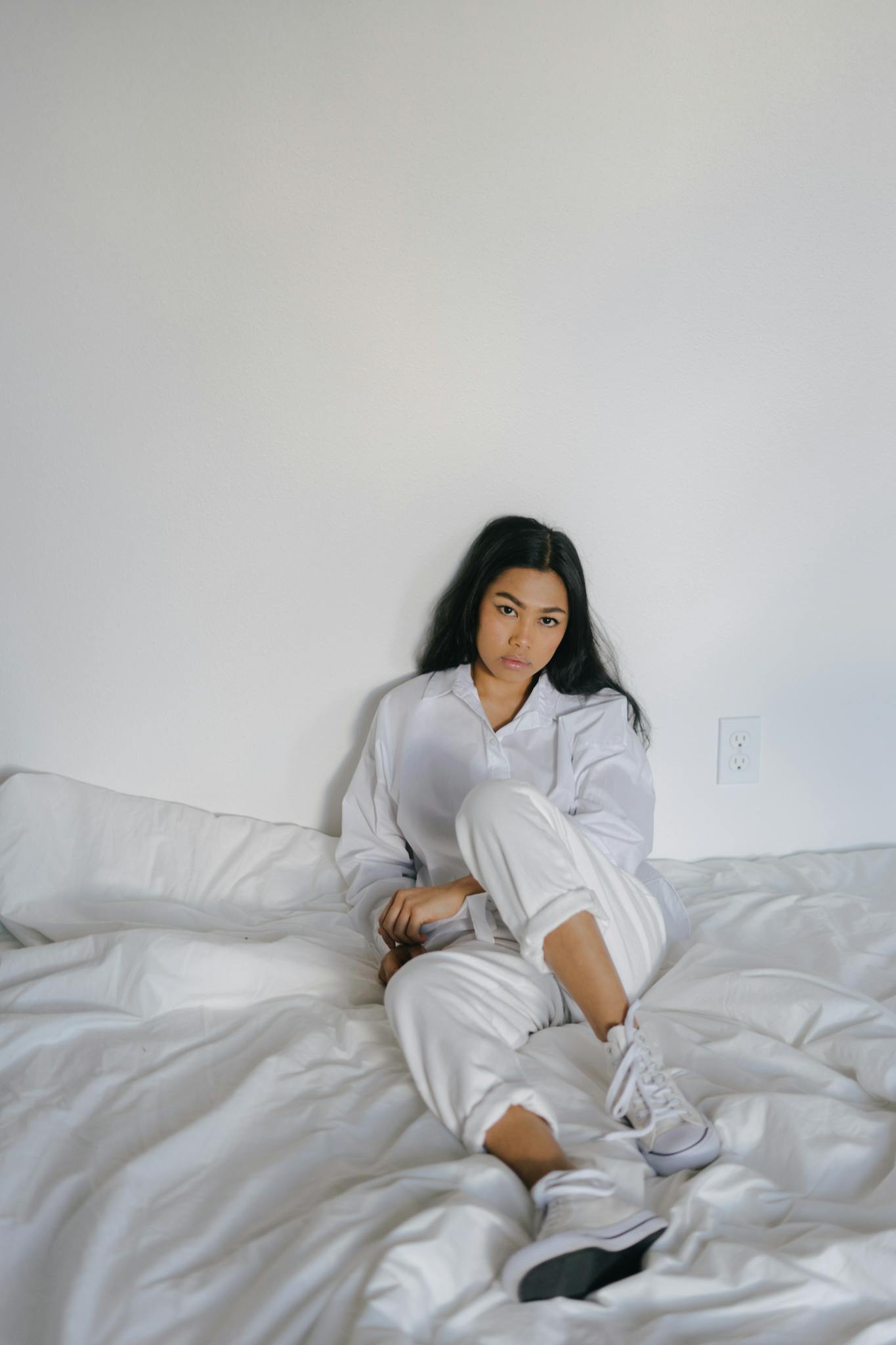 Casual portrait of a young woman sitting comfortably on a bed in white attire, indoors.