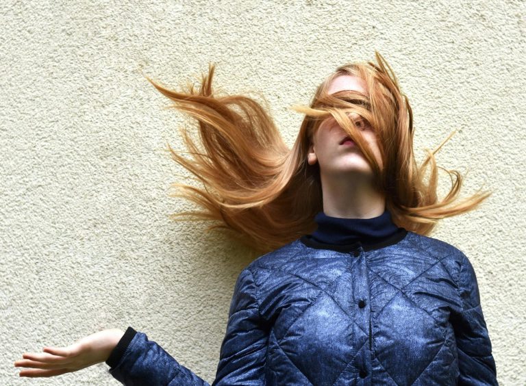 Stylish blonde woman with hair flipping in a bold pose against a textured wall.