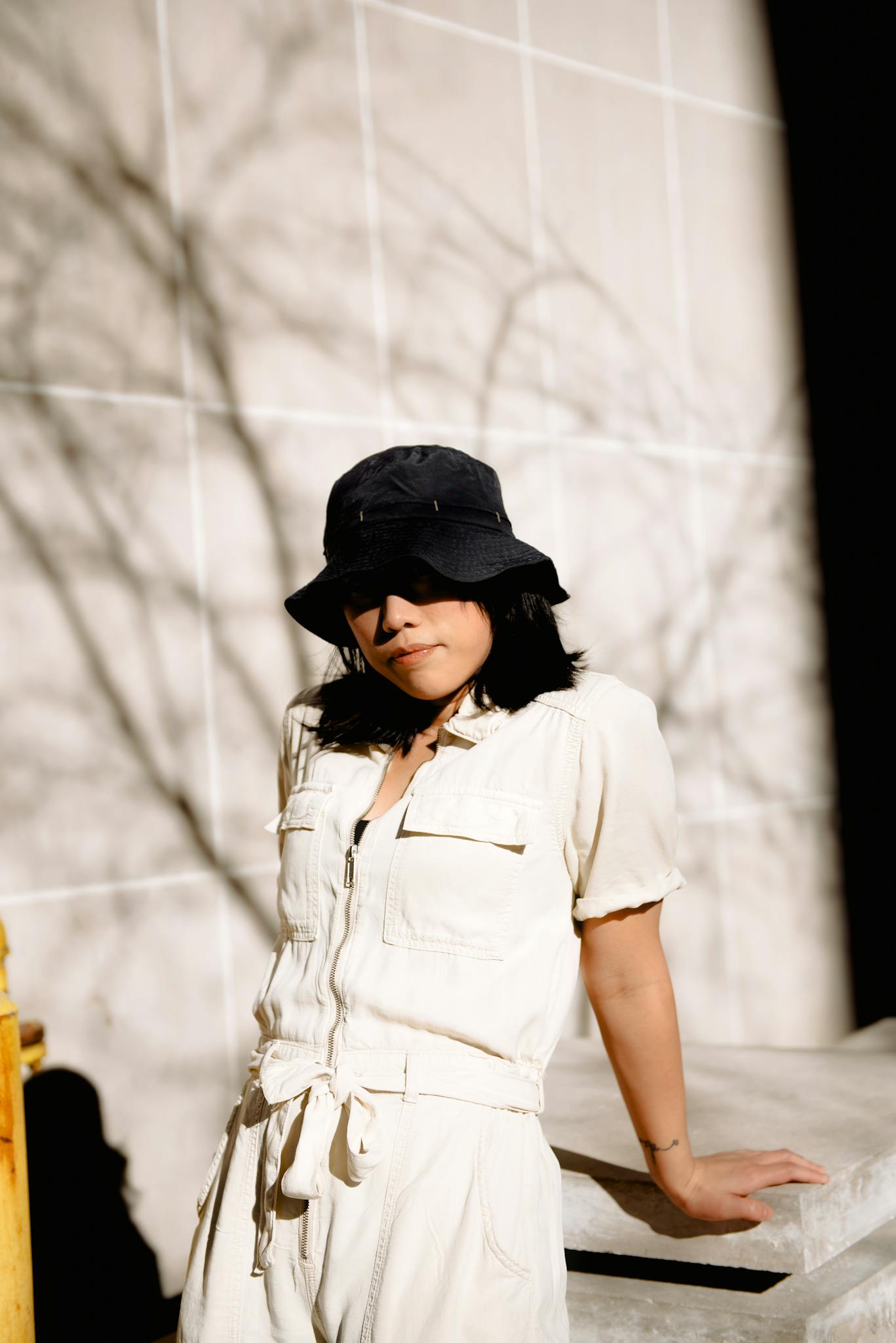 Woman in casual beige outfit and black hat leaning against sunlit wall outdoors.
